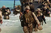 color photo of two columns of Marines wade through waist deep water disembarking from a landing craft onto a beach