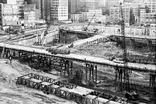 1968年,世贸大厦的基坑挖掘状况。中央是一个大坑,坑底有土,土上建造有桥梁,坑中有三台高耸的起重机,坑外是密集的高楼大厦。