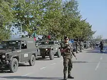 一名士兵站在一列軍事車隊前,車隊上寫著KFOR(科索沃維和部隊),並別著葡萄牙的國旗。第二輛車為葡萄牙的VBL。