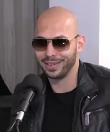 A bald man with a beard and sunglasses smiles at something off-camera with a microphone near his face.