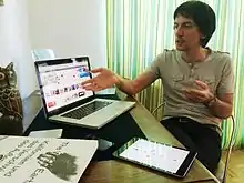 右側有一身穿茶色上衣和黑色褲子的黑髮男性,左側為他在展示《Blek》登上App Store官網的應用程式橫幅,圖片下方為正開啟《Blek》的iPad