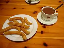 画面右侧是一杯浓稠、微微冒泡的热巧克力,一个不锈钢勺子插入巧克力中,杯子放置在杯垫上,杯身写着“ITAL CAFFÈ”。左侧是一盘六根交叠、弯曲的西班牙油条,每根呈长条状,表面有顺着长边的凹槽。