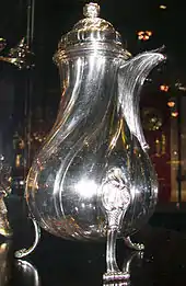 一个光滑的金属巧克力壶,上窄下粗。顶部是半球形的盖子,刻有凹槽,右侧有壶嘴,底部是三个向外弯曲的支撑,刻有繁复的花纹。壶放置于博物馆中。