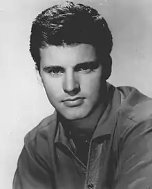 A young man in profile playing a guitar and standing before a microphone.