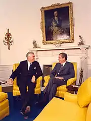 Two men sit in golden-yellow chairs and appear to be conversing cordially.