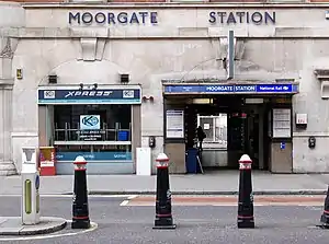 A grey building with a rectangular, dark blue sign reading "MOORGATE STATION" in white letters and short, black, white-tipped posts in front