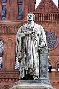 An aged bronze statue of a man wearing a robe. His left hand rests on a book which sits on a pedestal and the statue is in front of a red brick building reminiscent of a Romanesque Cathedral