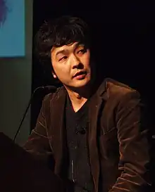 一名站在讲台上,身着棕色夹克和深灰衬衫的日本男子