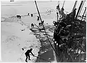 Men with digging tools removing ice surrounding the ships hull, creating an icy pool of water